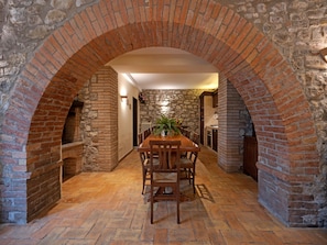 Um corredor de tijolos com arco que leva a uma área de refeições, com mesa e cadeiras, paredes de pedra e chão em mosaico.
