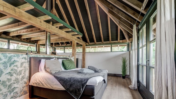 Una camera da letto con soffitto in legno, un letto con cuscini, una pianta in vaso e una finestra con tende.