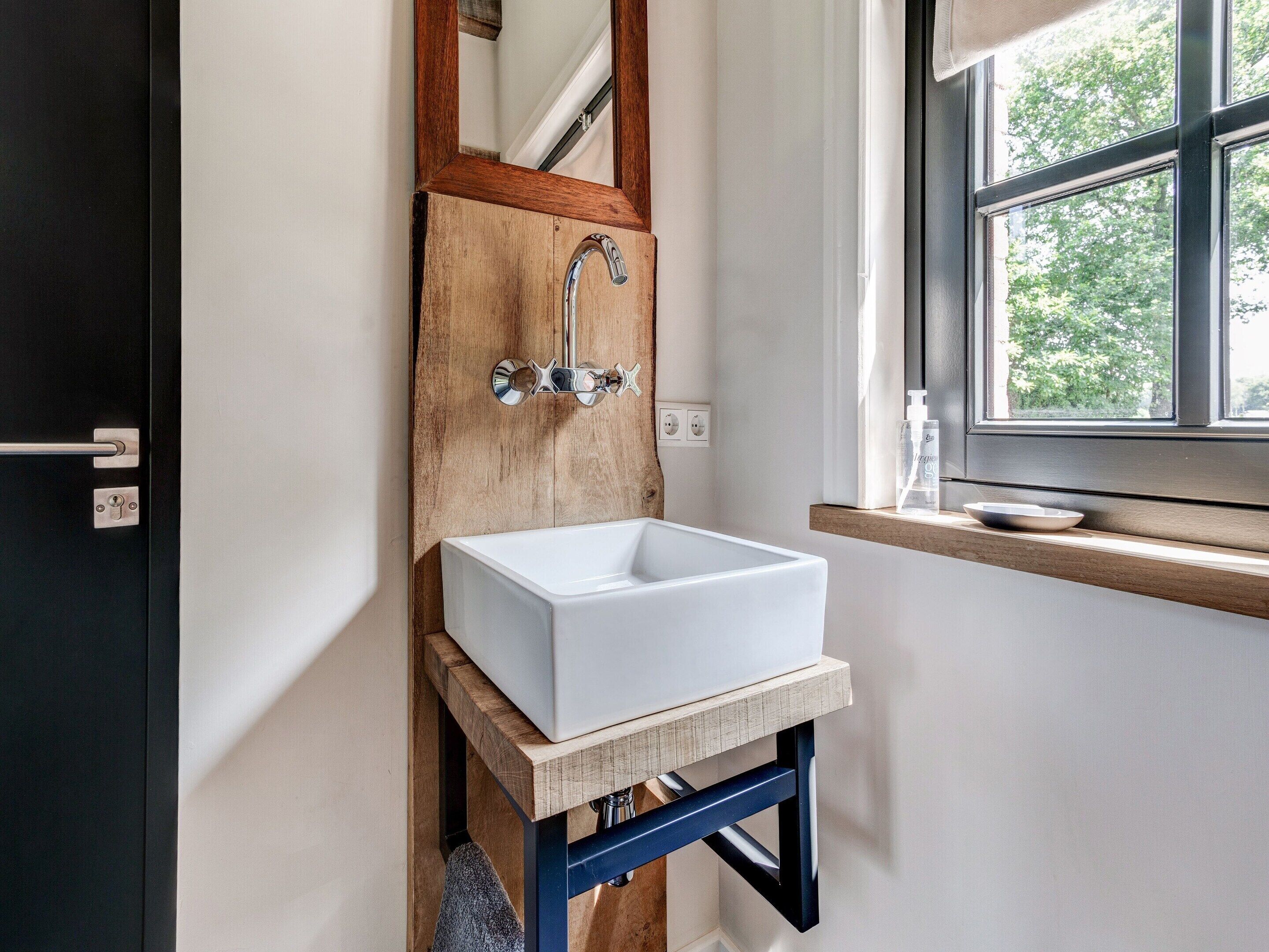 A bathroom with a white sink mounted on a wooden shelf, a wooden-framed mirror above the sink, and a window with a view of trees.