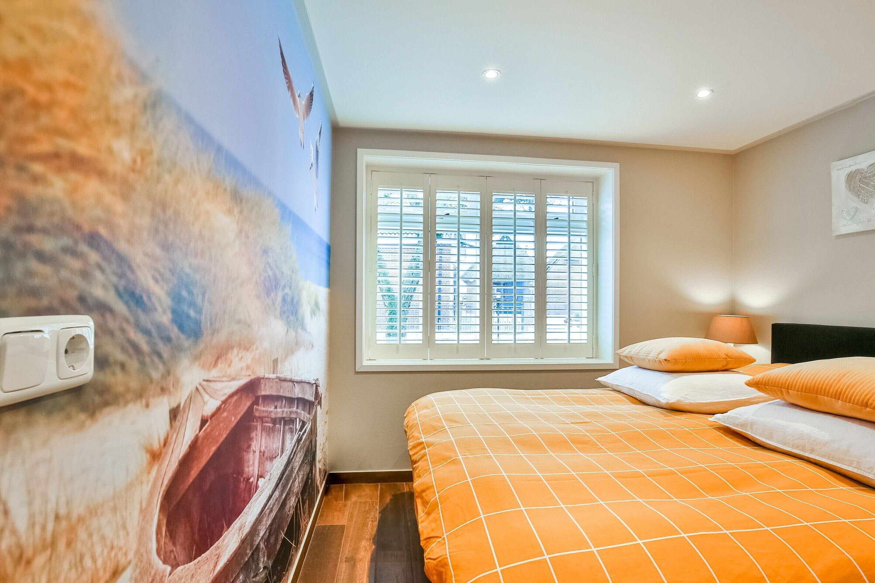 A bedroom with a mural of a beach scene, a window with blinds, an orange bedspread, and a bedside lamp.