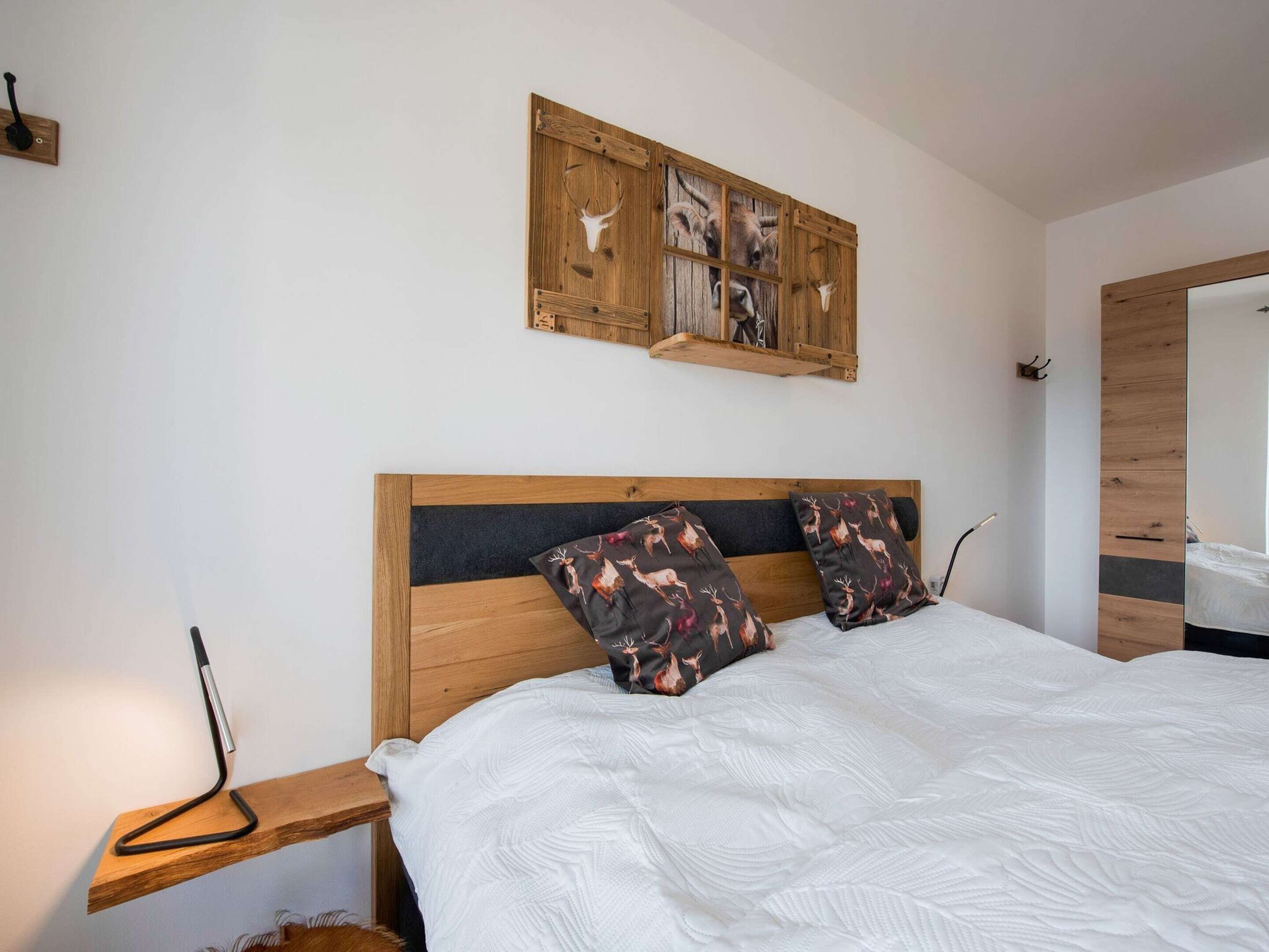Un dormitorio con cabecera de madera, colcha blanca, dos almohadas con estampado de ciervo y una repisa de madera con una lámpara.