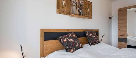 Un dormitorio con cabecera de madera, colcha blanca, dos almohadas con estampado de ciervo y una repisa de madera con una lámpara.