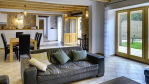 A spacious living area with a leather sofa, dining table, and chairs. A kitchen area is visible in the background with stone walls and wooden beams. - Villa in Batlug With Private Pool (Gracisce)