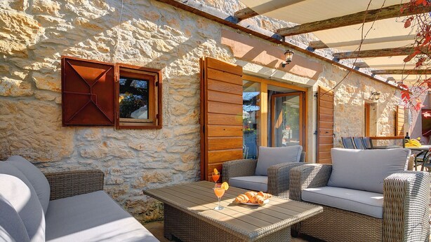 A stone-walled outdoor seating area with wicker furniture and a table, featuring a glass door and wooden beams. - Villa in Batlug With Private Pool (Gracisce)