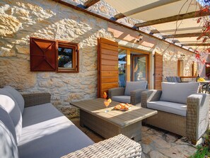 Un espace extérieur aménagé avec des murs en pierre, doté de meubles en osier et d’une table, et agrémenté d’une porte vitrée et de poutres en bois.