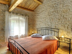 Une chambre avec un mur en pierre, un lit avec un cadre en métal, un plafond en bois et une fenêtre ornée de rideaux légers.