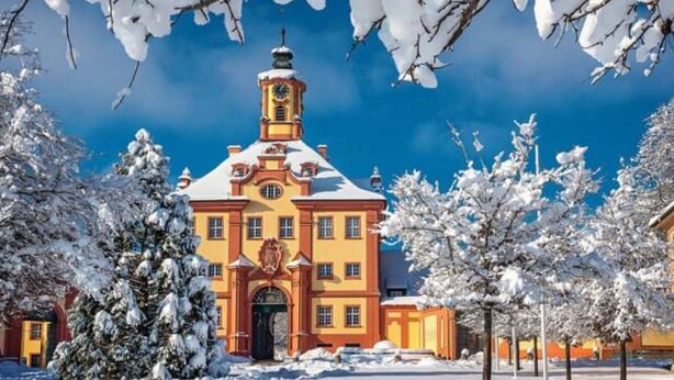 Ein großes, gelbes Gebäude mit einem markanten Turm und einer Uhr, umgeben von schneebedeckten Bäumen.