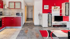Eine moderne Küche mit roten Schränken, einer Mikrowelle und einem Spülbecken. Ein Flur führt in einen Wohnbereich mit Fernseher und einer Glastruhe.