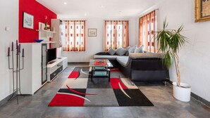 Ein modernes Wohnzimmer mit Sofa, Couchtisch und Fernseher. Es gibt eine Küchenzeile mit Kühlschrank und Spüle. Der Raum hat große Fenster mit Vorhängen.