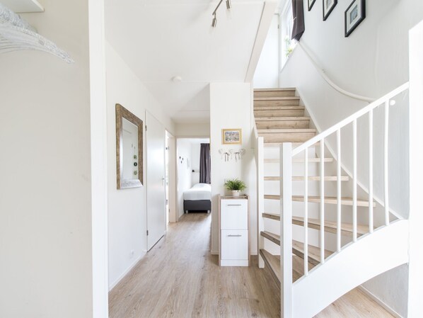 Un couloir avec un escalier, un plancher en bois, des murs blancs et une petite table de chevet sur laquelle est posée une plante.