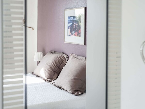 Een slaapkamer met een bed, kussens, een bedlampje en een ingelijst beeld aan de muur.