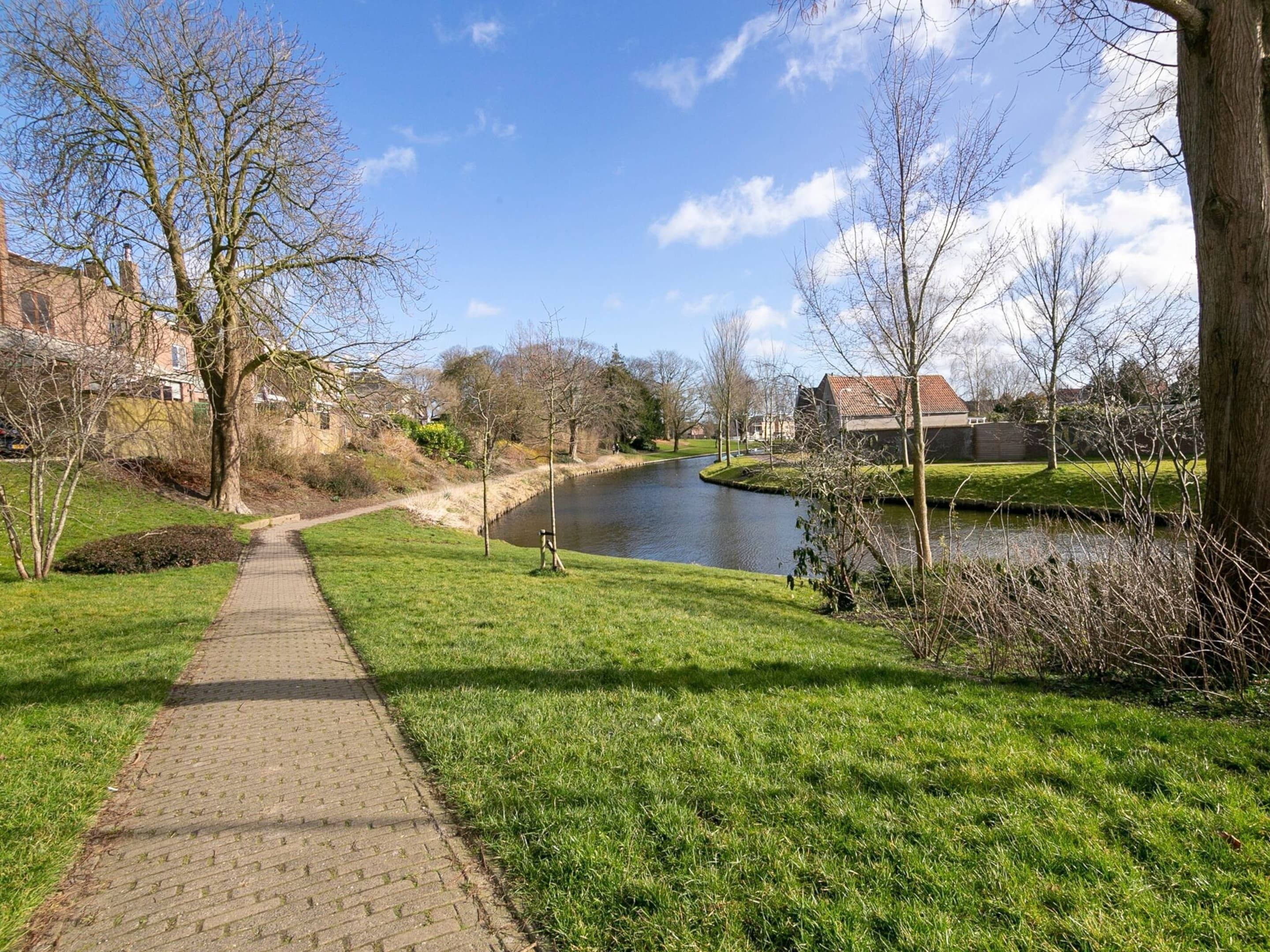 A paved pathway alongside a river, flanked by trees and buildings.