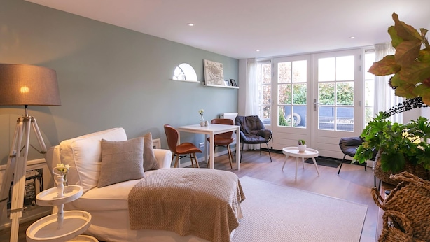 A cozy living room with a sofa, a coffee table, and a dining area with chairs. There is a window with a view of greenery outside. - Holiday Home in Medemblik With Garden (Medemblik)