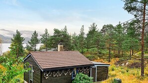 A small cabin with a tiled roof, surrounded by trees and a lake in the background. - 6 Person Holiday Home in Ostereidet (Alver)