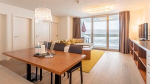 Ein modernes Hotelzimmer mit einem Holztisch, einer Couch mit Kissen, einem Fernseher und einer großen Glasschiebetür, die auf einen Balkon mit Meerblick führt.