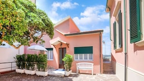 A pink building with a stone-paved courtyard, a white bench, and a tree providing shade. - Holiday Gome in San Vincenzo Near Beach (San Vincenzo)