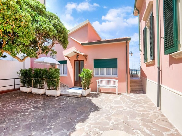 A pink building with a stone-paved courtyard, a white bench, and a tree providing shade. - Holiday Gome in San Vincenzo Near Beach (San Vincenzo)