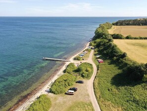 Eine Luftaufnahme einer Küstenlandschaft mit einem Steg, der sich ins Wasser erstreckt, einem Feldweg entlang des Ufers und geparkten Fahrzeugen.