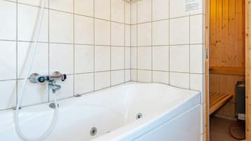 A white bathtub with a showerhead, tiled walls, and a wooden sauna visible through an open door.