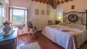A bedroom with a wooden ceiling, a bed with floral bedding, a chair, a table with a vase, and a window with a view. - Tuscany Bliss: Garden Retreat (San Casciano in Val di Pesa)