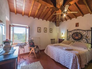 Un dormitorio con techo de madera, una cama con ropa de cama floral, una silla, una mesa con un jarrón y una ventana con vistas.