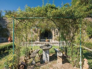 Un jardín con un pérgola, bancos de piedra, un abrevadero para pájaros y un rústico muro de piedra.