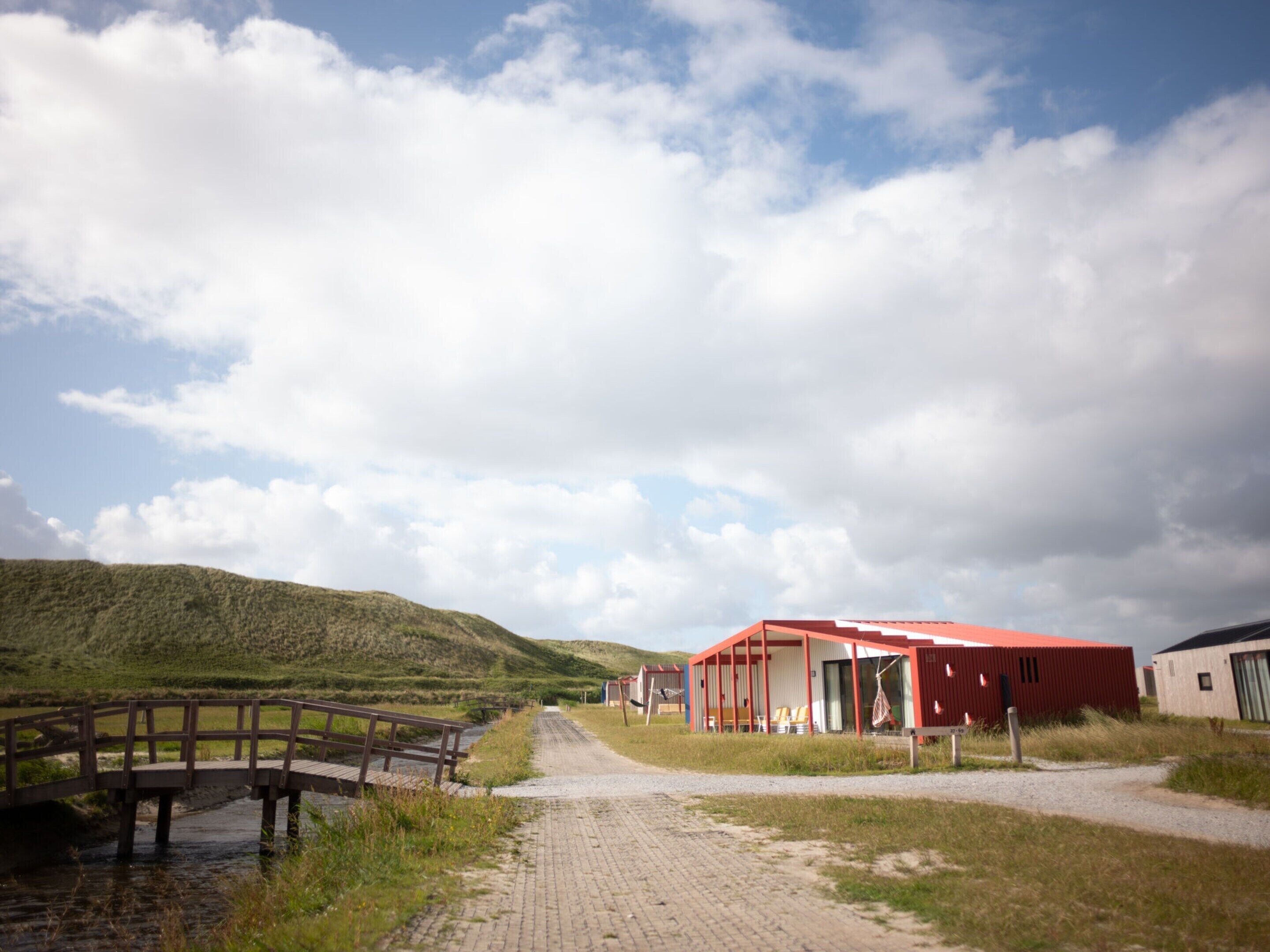 Un sentier mène à un bâtiment à toit rouge, doté d’un espace extérieur abrité, d’un pont en bois qui enjambe un ruisseau et, au loin, d’une colline verdoyante.