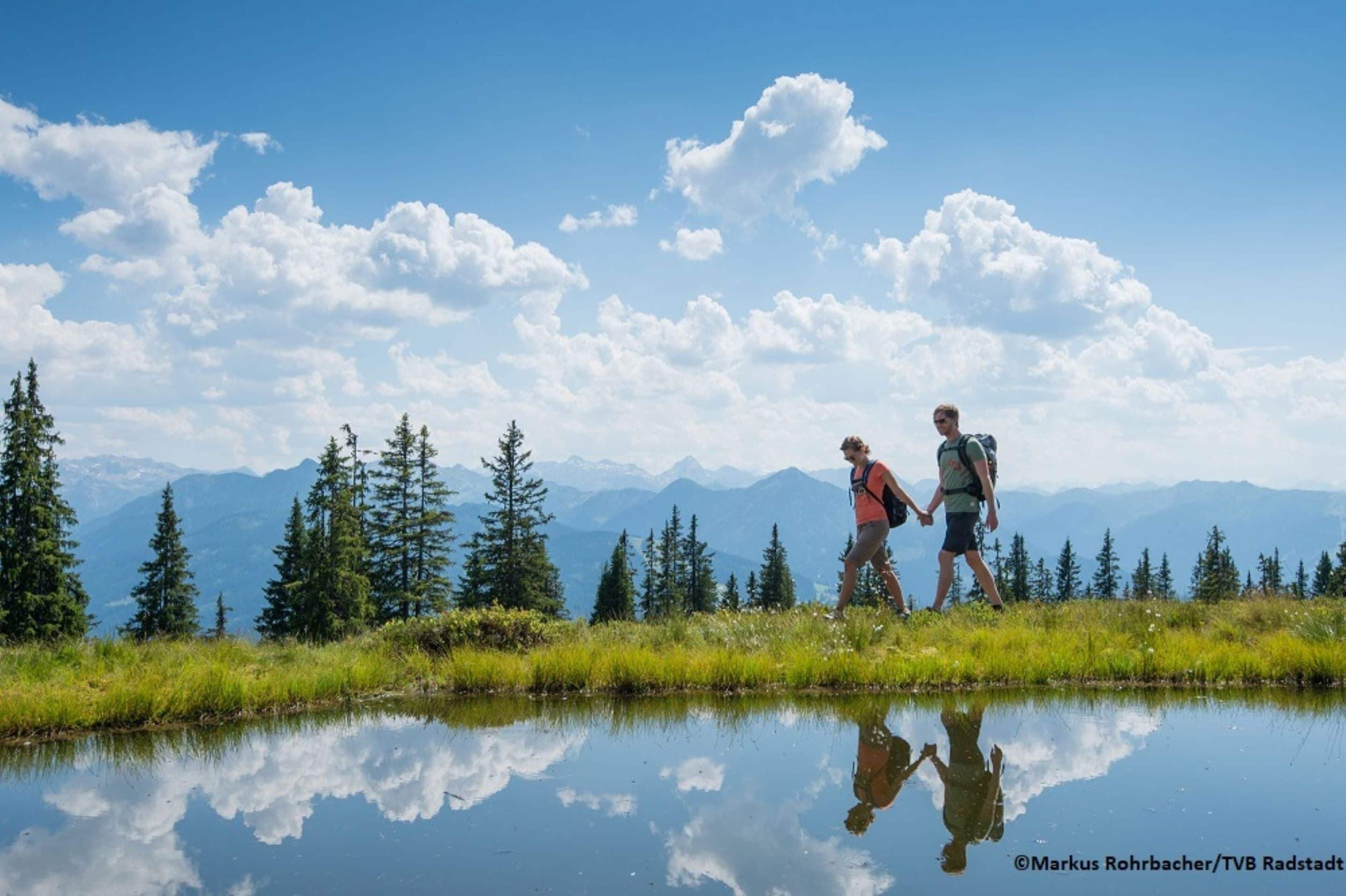 Two people walking on a grassy field with a body of water in the foreground and mountains in the background.