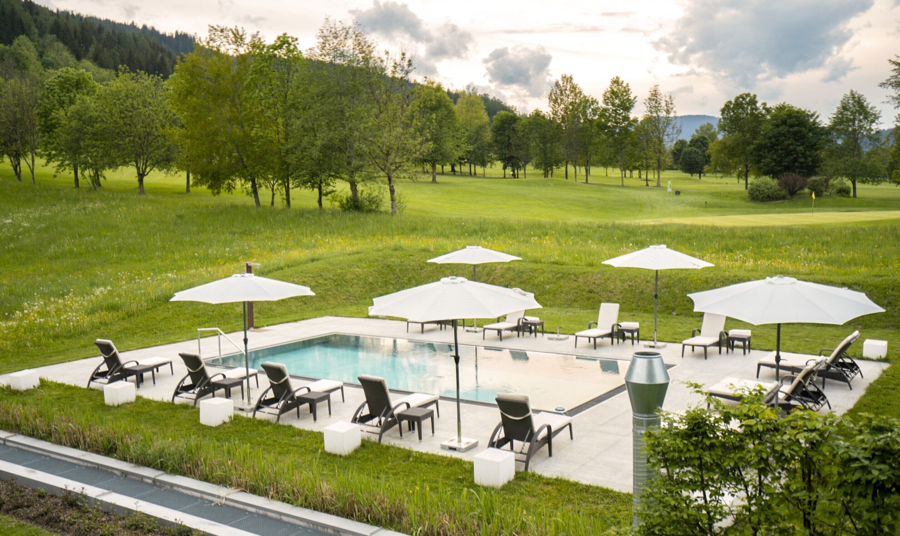 A poolside area with lounge chairs and umbrellas, surrounded by a lush green landscape and trees.