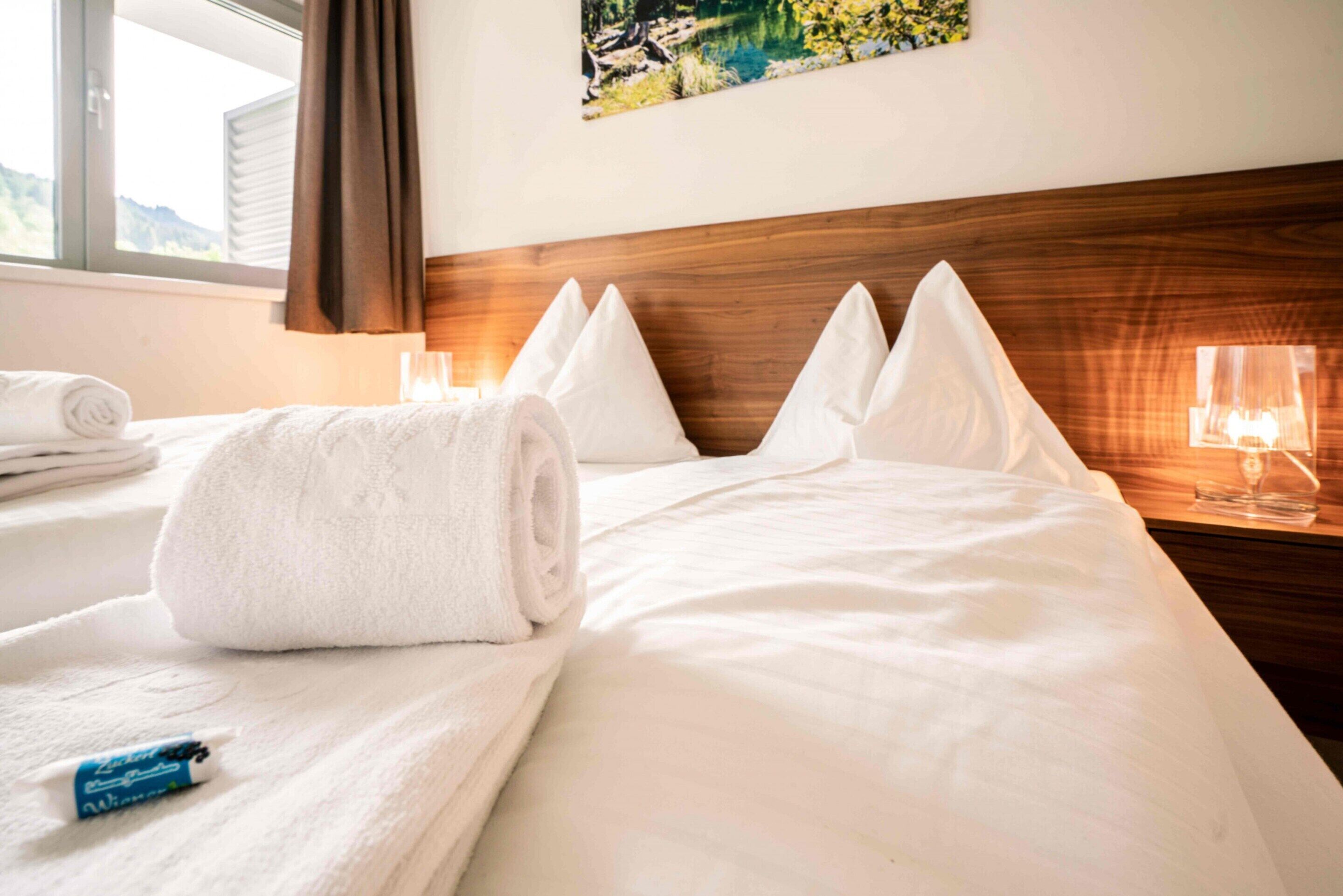 A hotel room with a neatly made bed, a rolled white towel, and a bedside table with a glass and a candle.