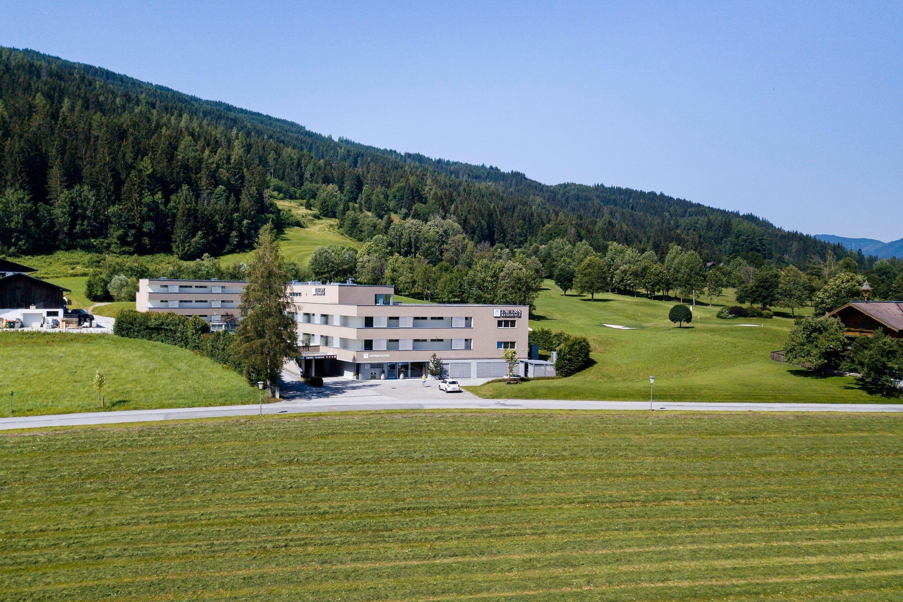 A modern building with a flat roof, surrounded by a well-maintained lawn and a forested hillside.