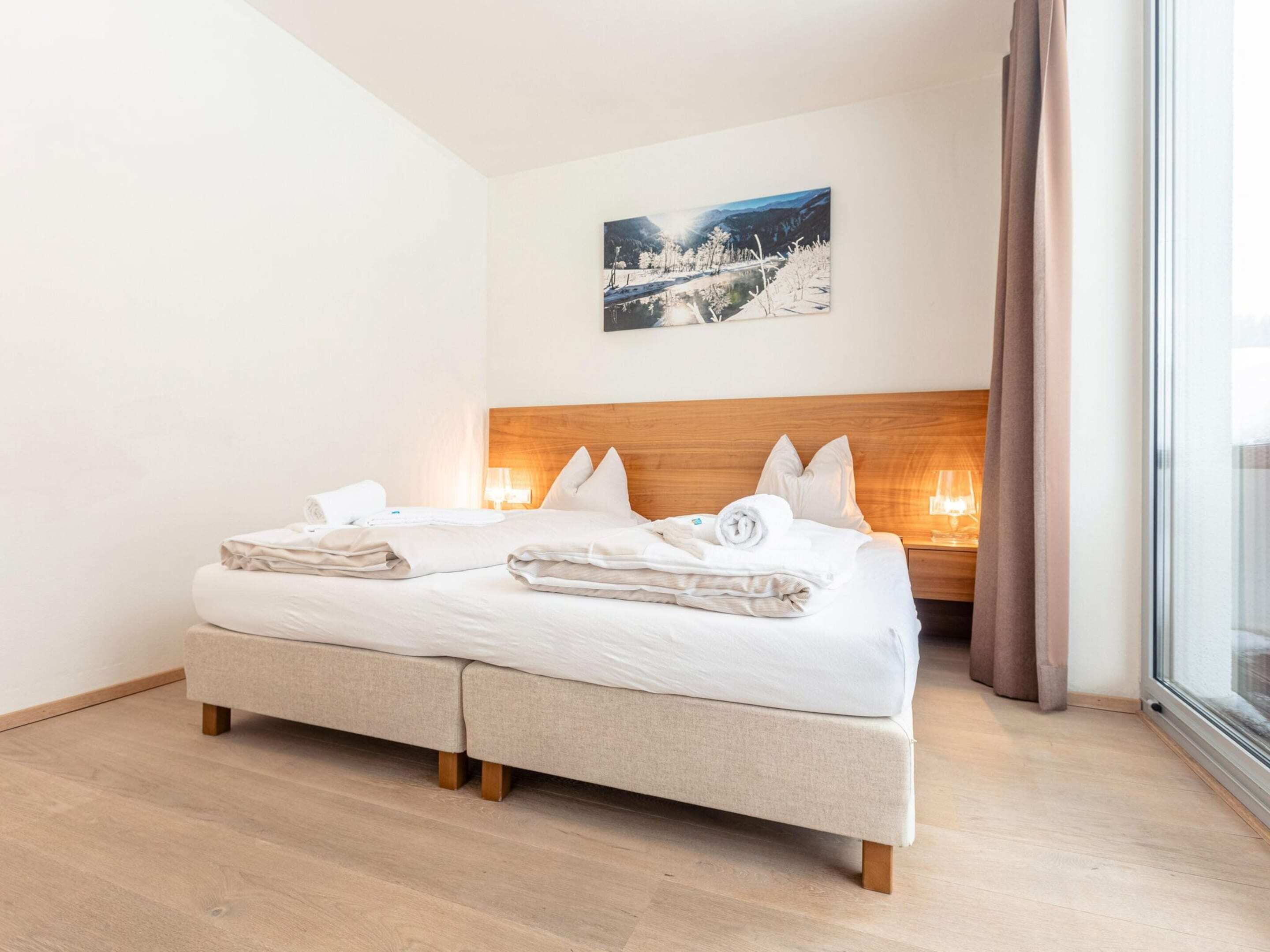 A double bed with a wooden headboard, two bedside tables with lamps, a painting on the wall, and a large window with curtains.