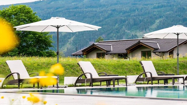 A pool area with lounge chairs and umbrellas, set against a backdrop of a mountain and a building with a sloped roof. - Nice Apartment With Dishwasher, Near the Ski Slopes (Radstadt)