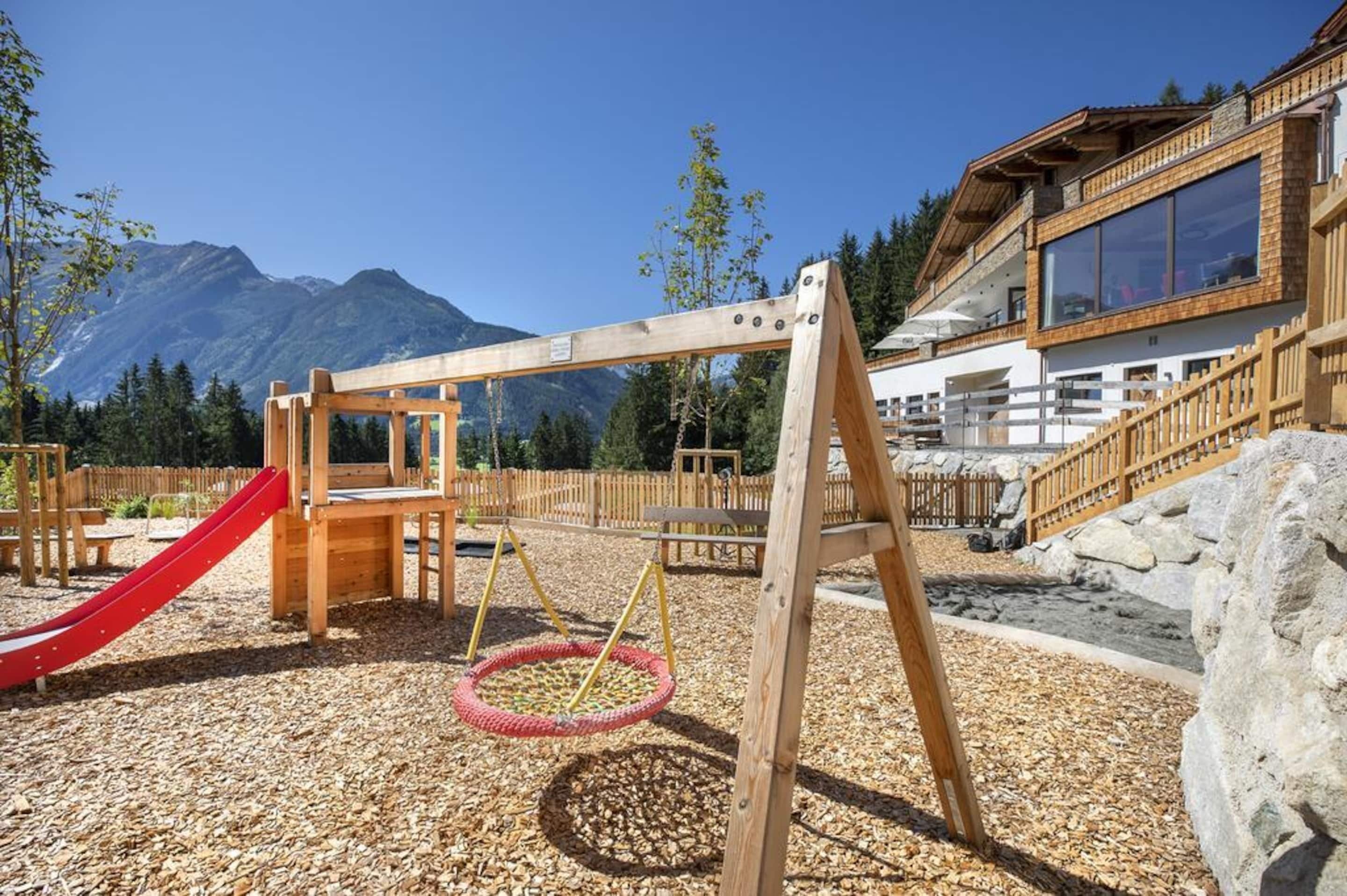 A wooden swing set and a red slide in a playground area with a mountain backdrop.