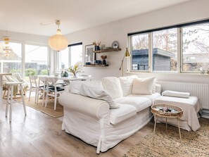 A modern living room with a white sofa, dining table, and chairs, a large window with a view, and a hanging light fixture. - 6 Person Holiday Home in Rude (Bisserup)