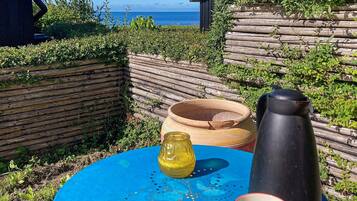 A blue table with a yellow pitcher, a black container, and a patterned cup, set against a backdrop of a wooden fence and a view of the sea.