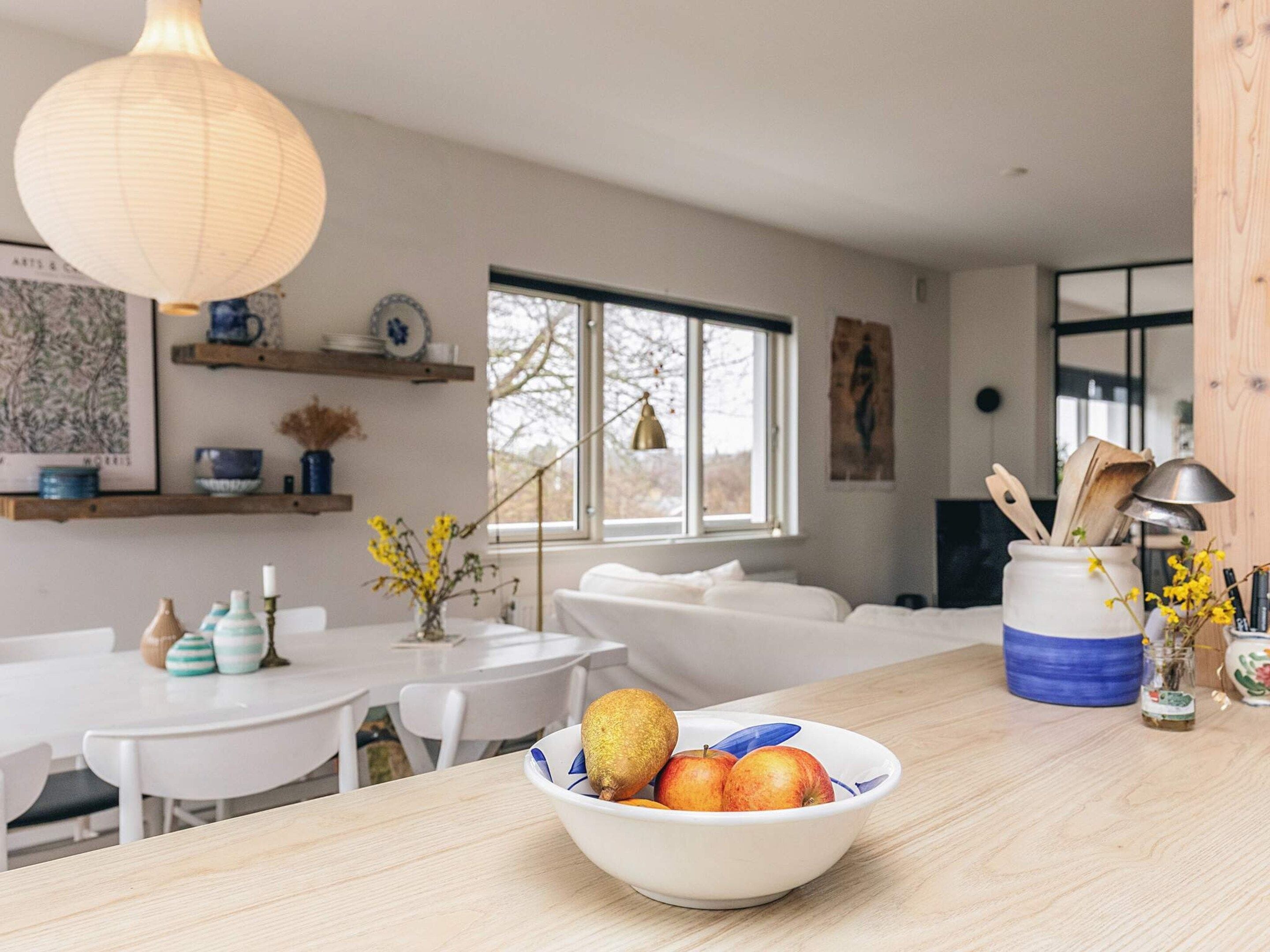A modern kitchen with white furniture, a bowl of fruit on the counter, and a large window.