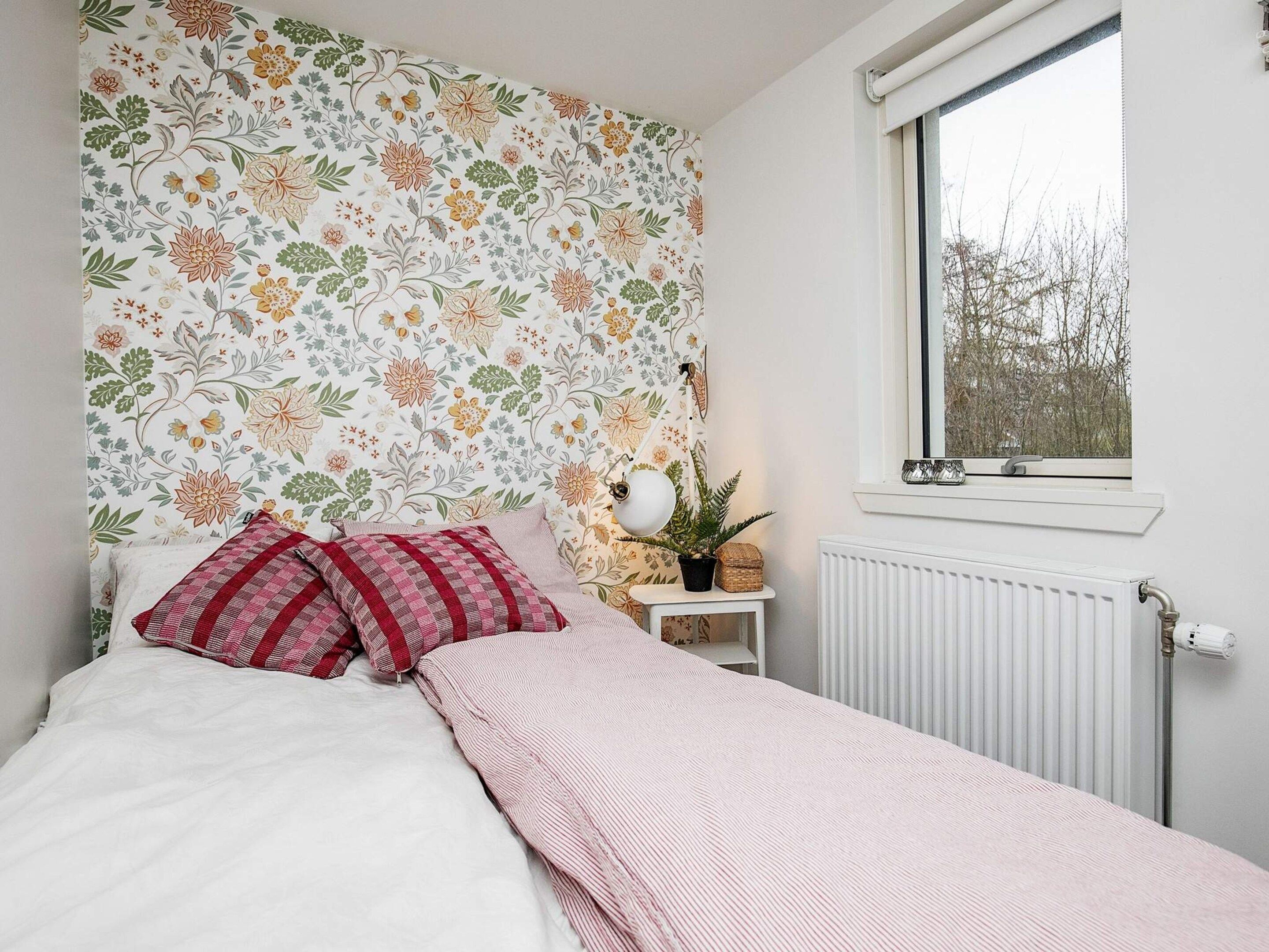 A bedroom with a floral wallpaper, a bed with white bedding and red pillows, a bedside table with a lamp and a plant, and a window with a view of trees.
