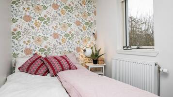 A bedroom with a floral wallpaper, a bed with white bedding and red pillows, a bedside table with a lamp and a plant, and a window with a view of trees.