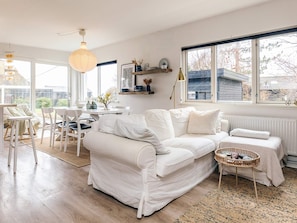 Ein modernes Wohnzimmer mit einem weißen Sofa, einem Esstisch, Stühlen, einem großen Fenster mit Aussicht und einer Hängeleuchte.