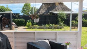 A covered outdoor seating area with a wicker chair and a small table, overlooking a garden with a wooden fence and a thatched roof structure. - 6 Person Holiday Home in Rude (Bisserup)