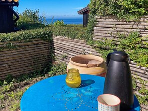 Uma mesa azul com um jarro amarelo, um recipiente preto e uma xícara estampada, com um cercado de madeira e vista para o mar ao fundo.