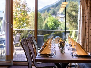 Une table à manger pour deux personnes, avec des chaises, donnant sur un paysage montagneux grâce à de grandes fenêtres, et un balcon.