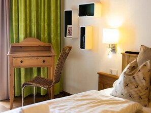 Een hotelkamer met een bed, een bureau met lades, een rieten stoel, een nachtkastje met een lamp en een groen gordijn.