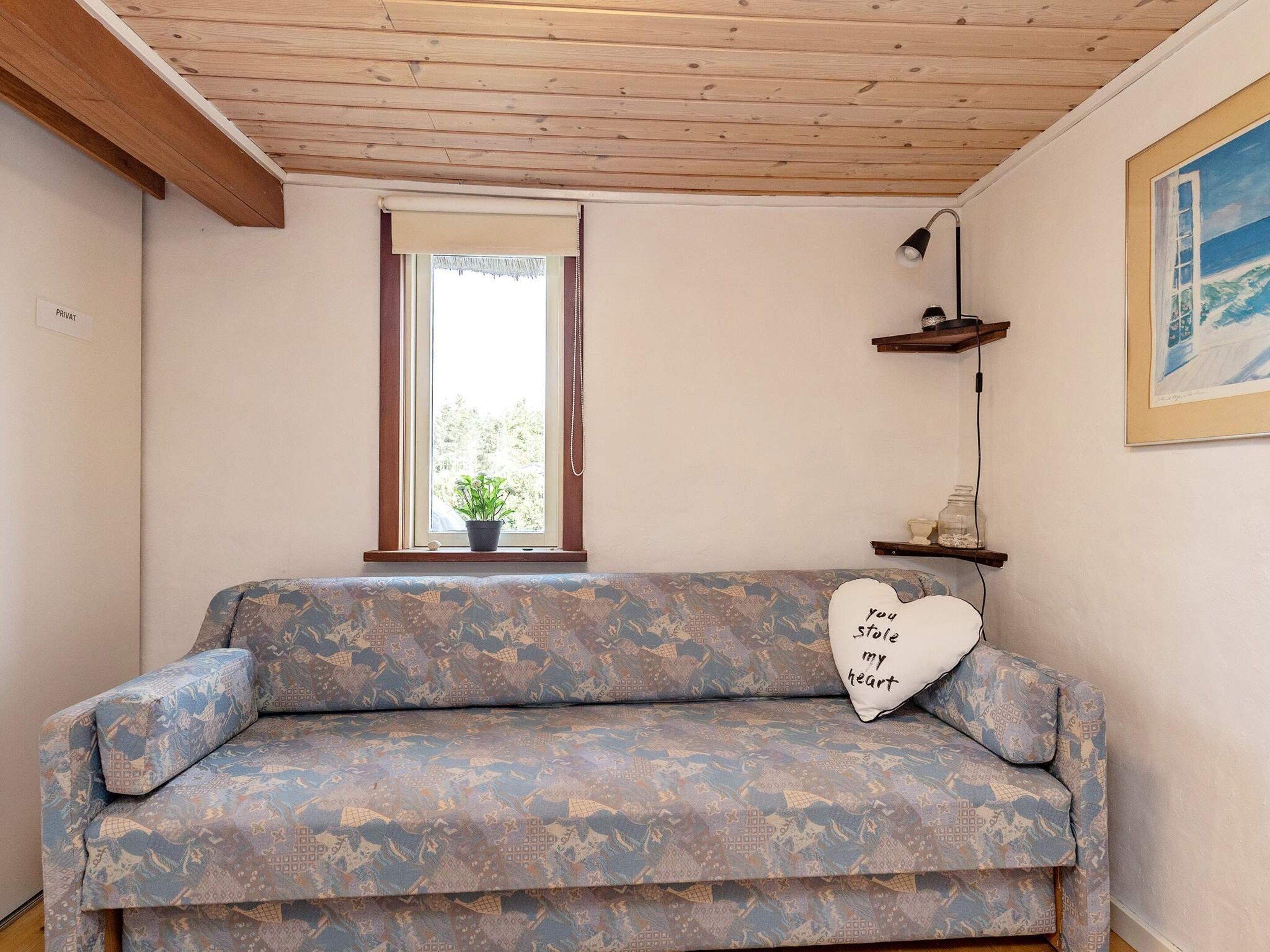 Una habitación con un sofá estampado, una ventana con una planta, un cojín con forma de corazón y un cuadro en la pared.