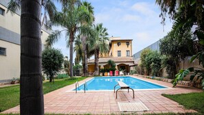 Una piscina con cubierta de madera, rodeada de palmeras y un edificio con techo de tejas.