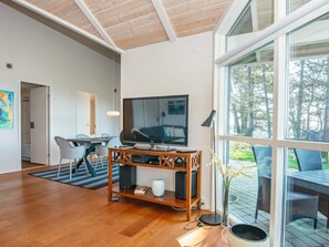 A modern living room with a large TV, wooden entertainment unit, and a dining area with chairs and a table. - Holiday Home in Ebeltoft (Ebeltoft)