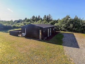A modern, single-story building with a dark exterior, a covered outdoor seating area, and a gravel driveway. - Holiday Home in Brovst (Brovst)