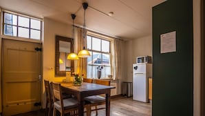 A compact kitchen with wooden flooring, a dining table with chairs, a hanging light fixture, a refrigerator, and a window with curtains. - Superb Holiday Home on a Mini Campsite Near the Beach (Vrouwenpolder)