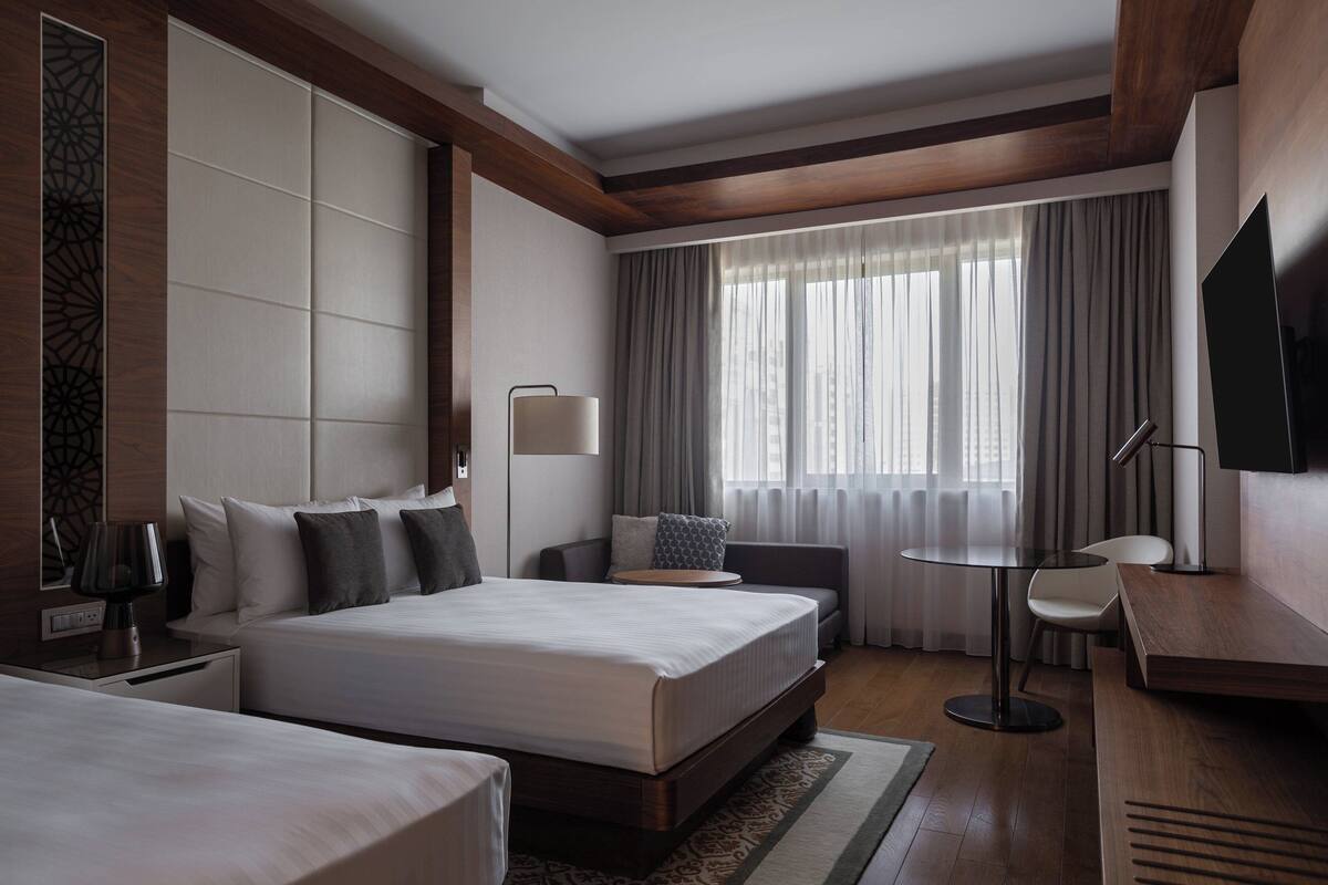 Habitación de hotel con dos camas, un escritorio, televisor y un ventanal con cortinas.